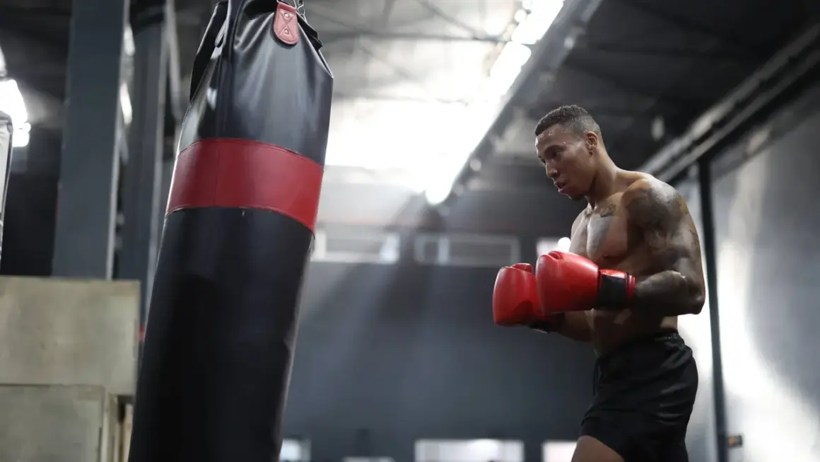 A boxer training intensely, sweat glistening on his skin as he throws punches at a heavy bag in a dimly lit gym.