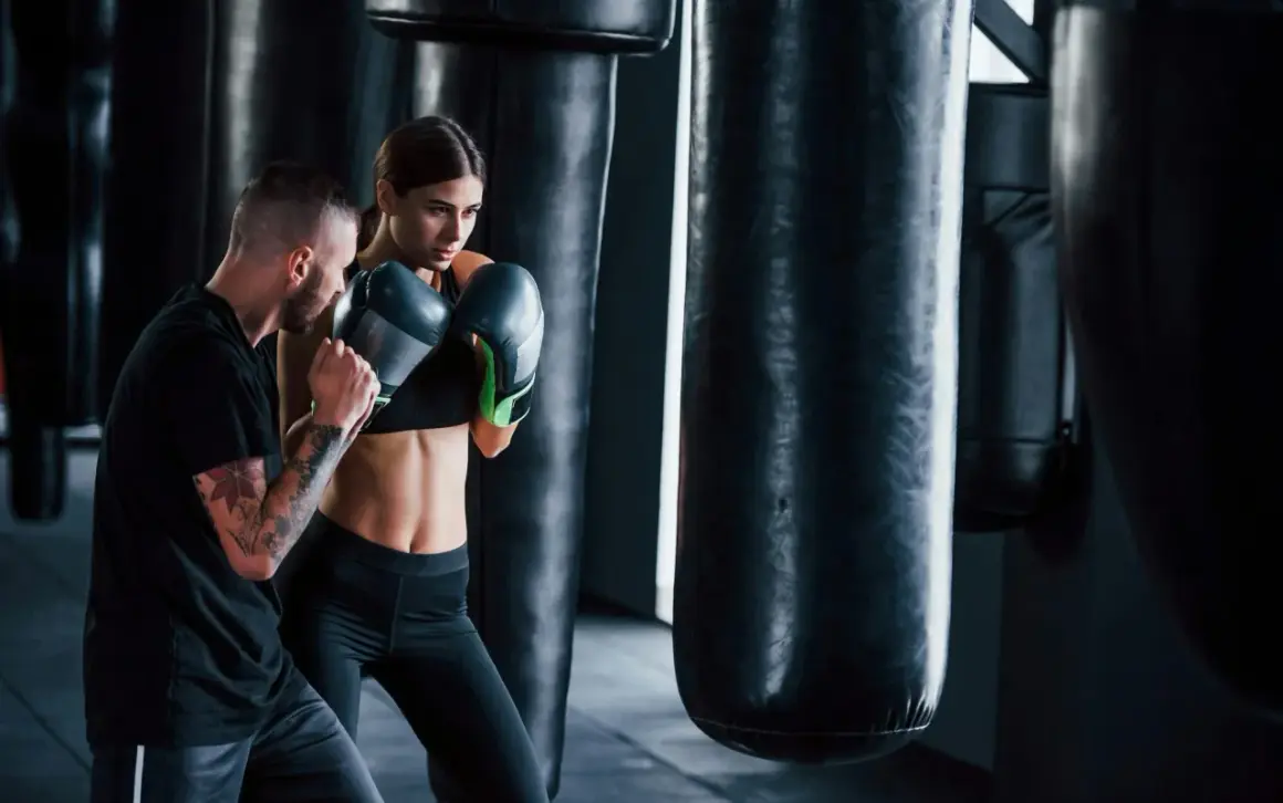A man with tattoos trains a woman in boxing gloves. They are in a gym with punching bags, showcasing the physical and mental boxing benefits.