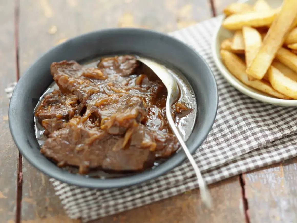 Un plat de carbonade flamande mijotée, servie dans un bol avec une cuillère, à côté d'une coupelle de frites dorées.