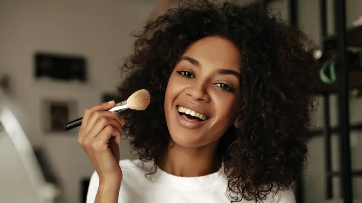 Donna sorridente con capelli ricci applica cipria con un pennello, mostrando come mettere la cipria senza pennello.