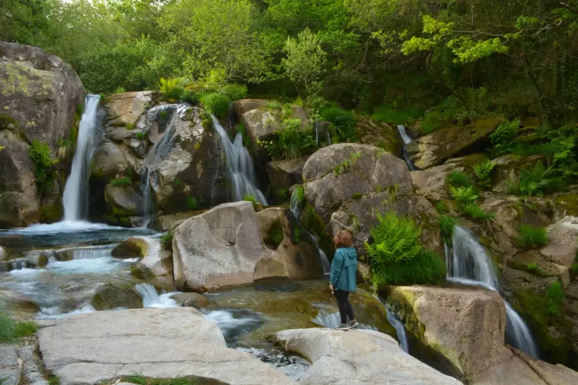 Una persona observa la hermosa fervenza de Noveira, con cascadas cayendo entre rocas cubiertas de musgo y vegetación exuberante.