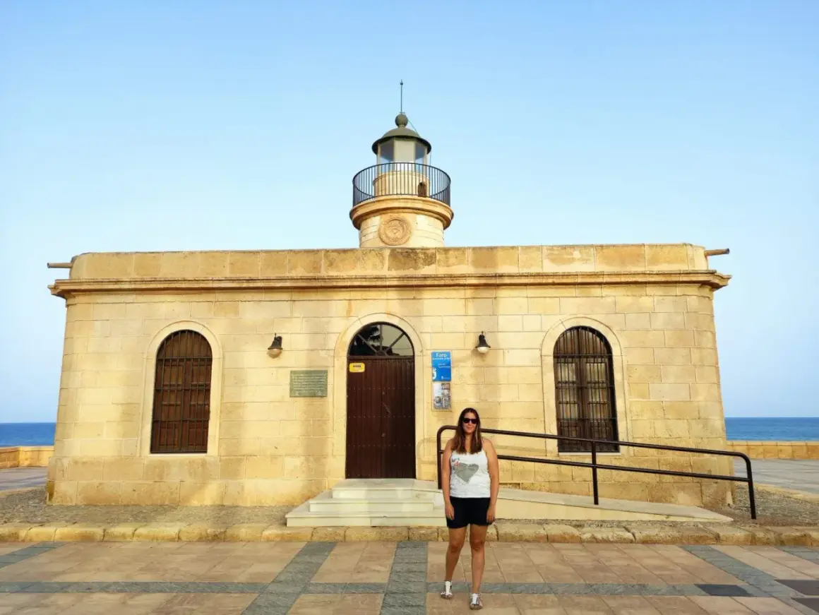Mujer sonriente posa frente al Faro de Roquetas de Mar, con el mar azul de fondo.