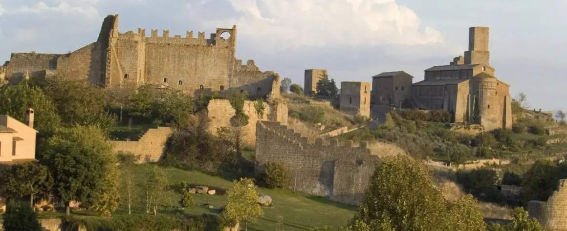 Antiche mura e torri di Tuscania VT si stagliano su una collina verdeggiante, con una chiesa imponente in lontananza.
