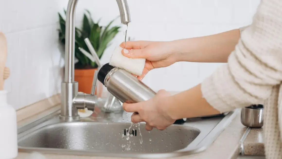 Hände reinigen eine Edelstahl-Thermoskanne unter fließendem Wasser im Spülbecken.