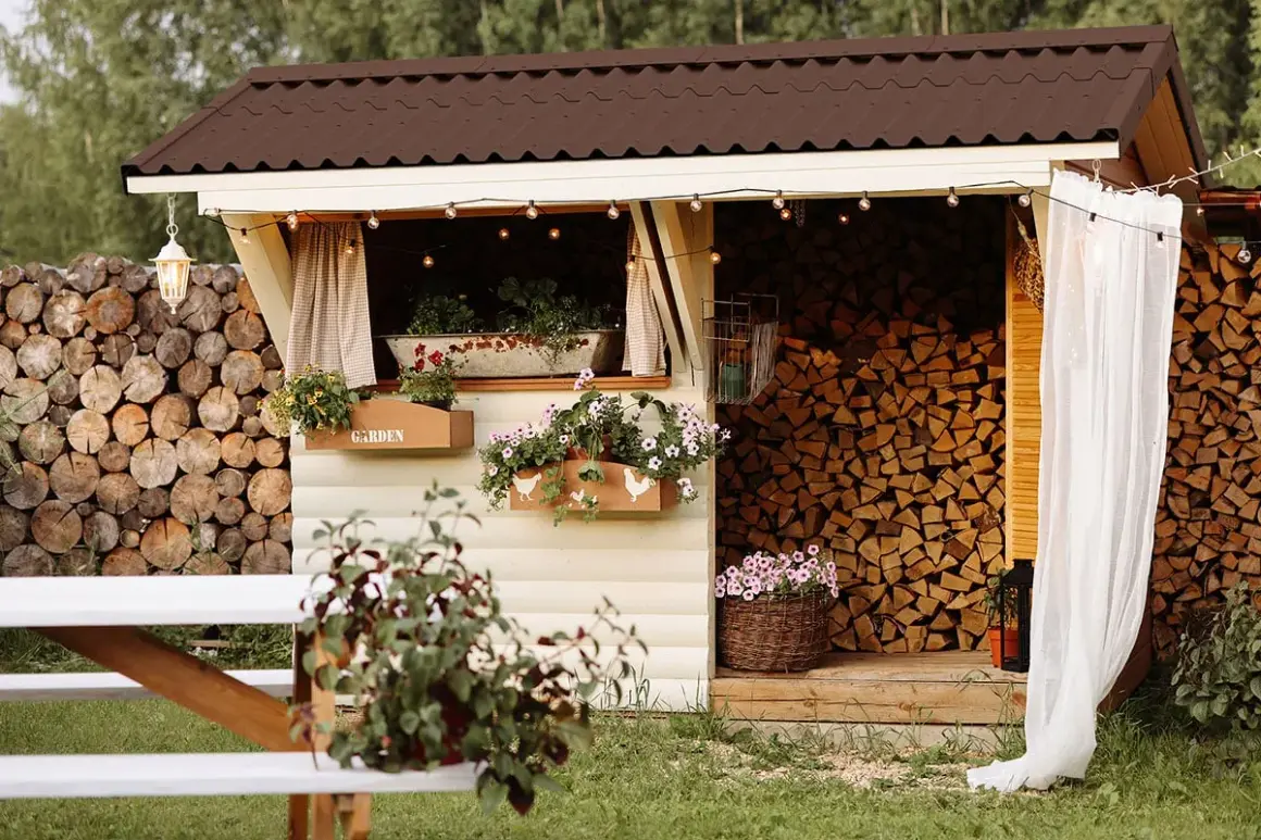 Charmante Holzunterstand Ideen: Ein gemütlicher Schuppen mit Blumenkästen, Lichterketten und einem Stapel Brennholz.