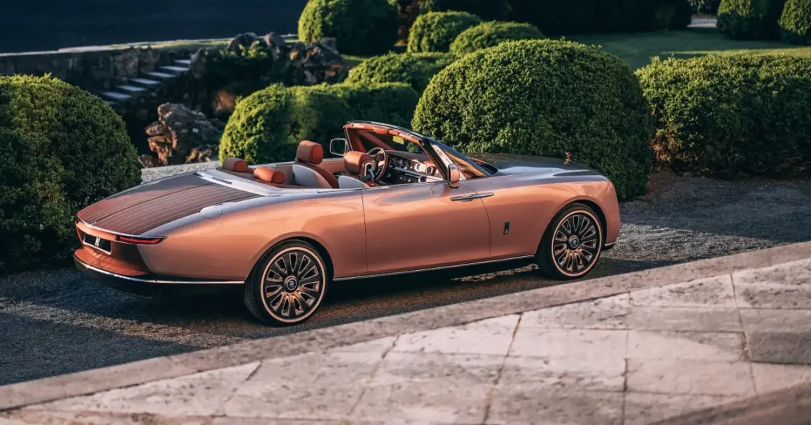 Elegancki Rolls-Royce Boat Tail w kolorze miedzianego różu, z drewnianym tyłem, zaparkowany wśród bujnej zieleni.