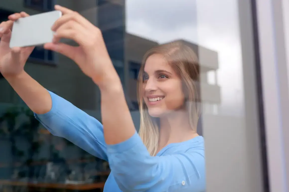 Uśmiechnięta blondynka robi sobie selfie w lustrze, trzymając telefon. Wskazówka, jak zrobić dobre selfie w lustrze: złap światło!