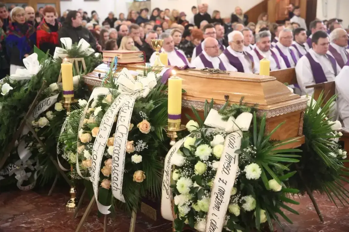 Ksiądz odczytuje ostatnie pożegnanie zmarłego przy trumnie, otoczonej wieńcami i świecami.