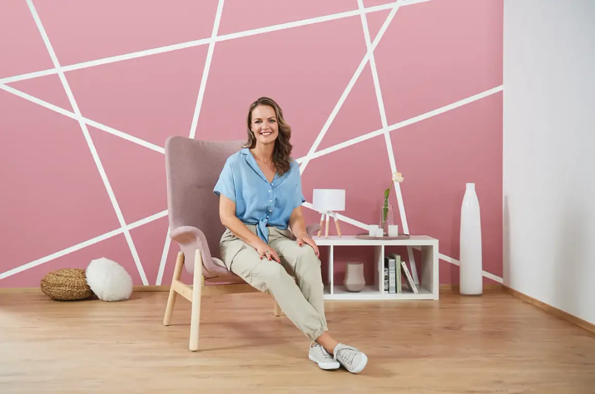 Frau sitzt vor einer Wandgestaltung mit Kreisen und Linien in Rosa und Weiß. Ein Sessel und ein Beistelltisch mit Deko stehen im Raum.