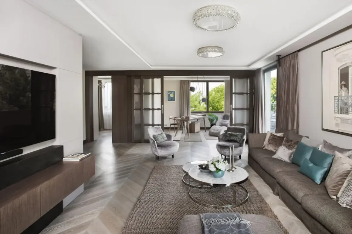 Elegancki salon z kanapą, fotelami i stolikiem kawowym. W tle widać jadalnię i okno z widokiem na zieleń. Całość emanuje wyrafinowanym, **włoskim stylem**.