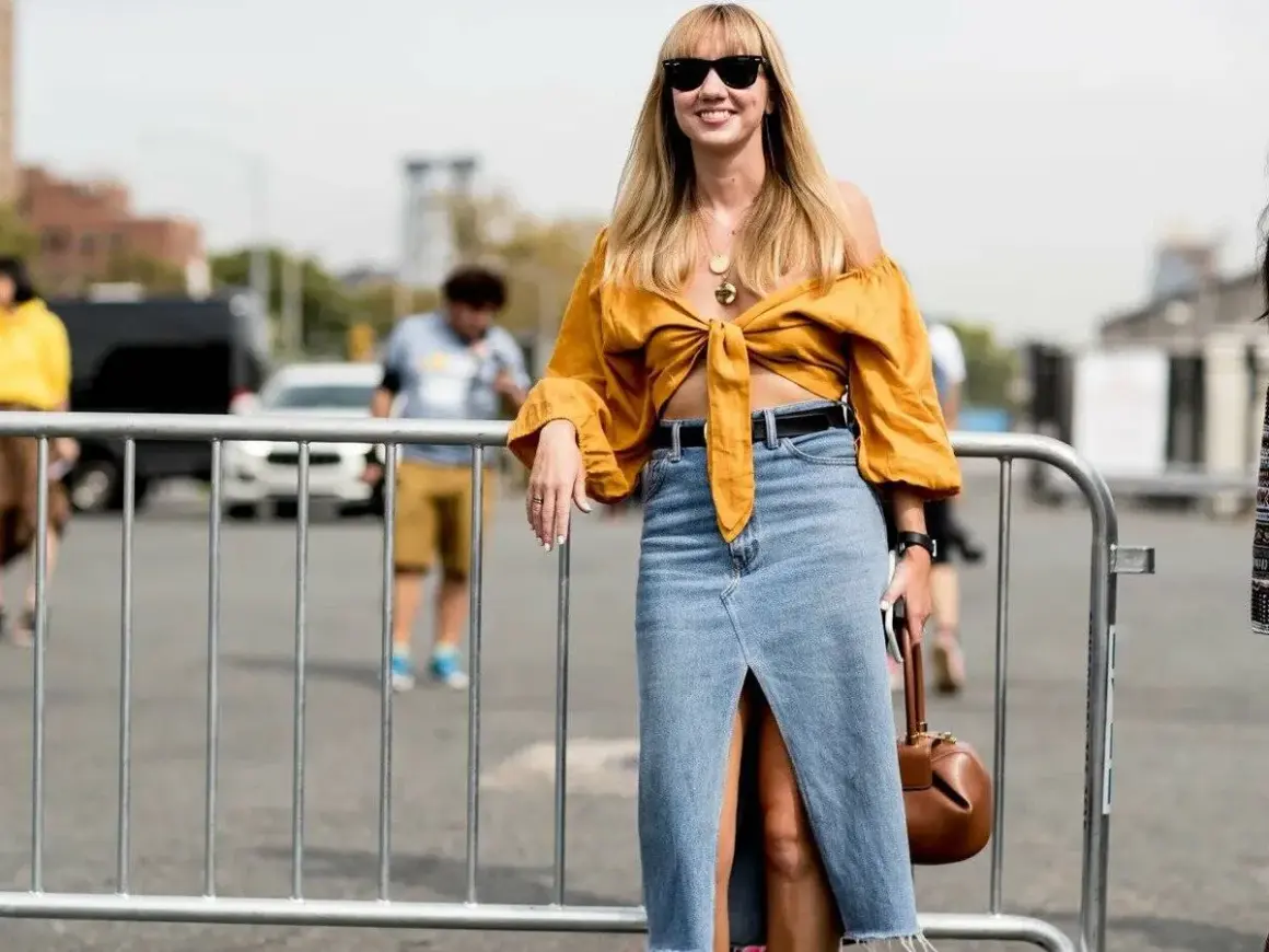 Mujer sonriente con un outfit falda vaquera larga, blusa amarilla anudada y bolso marrón.
