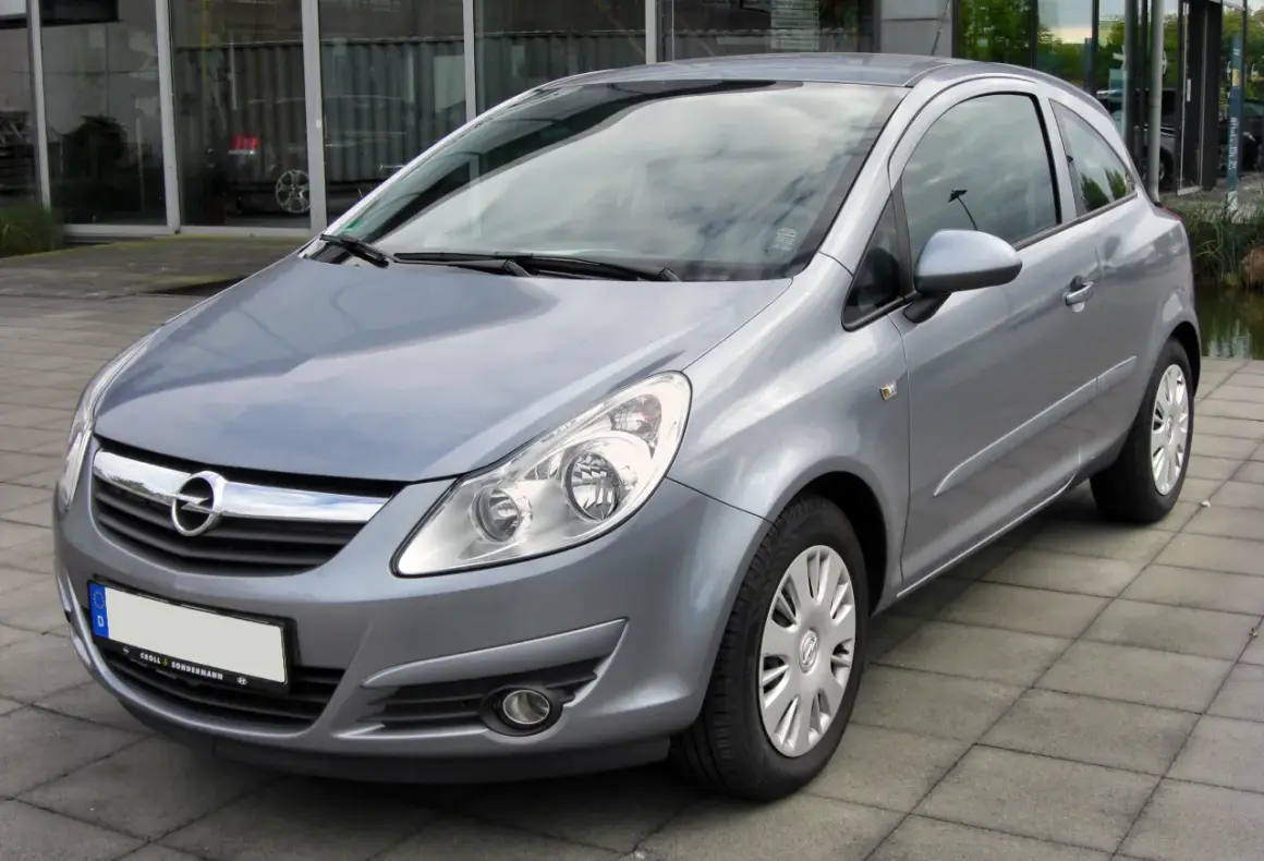 Srebrny Opel Corsa D 1.4 benzyna stoi na chodniku przed budynkiem z dużymi oknami.