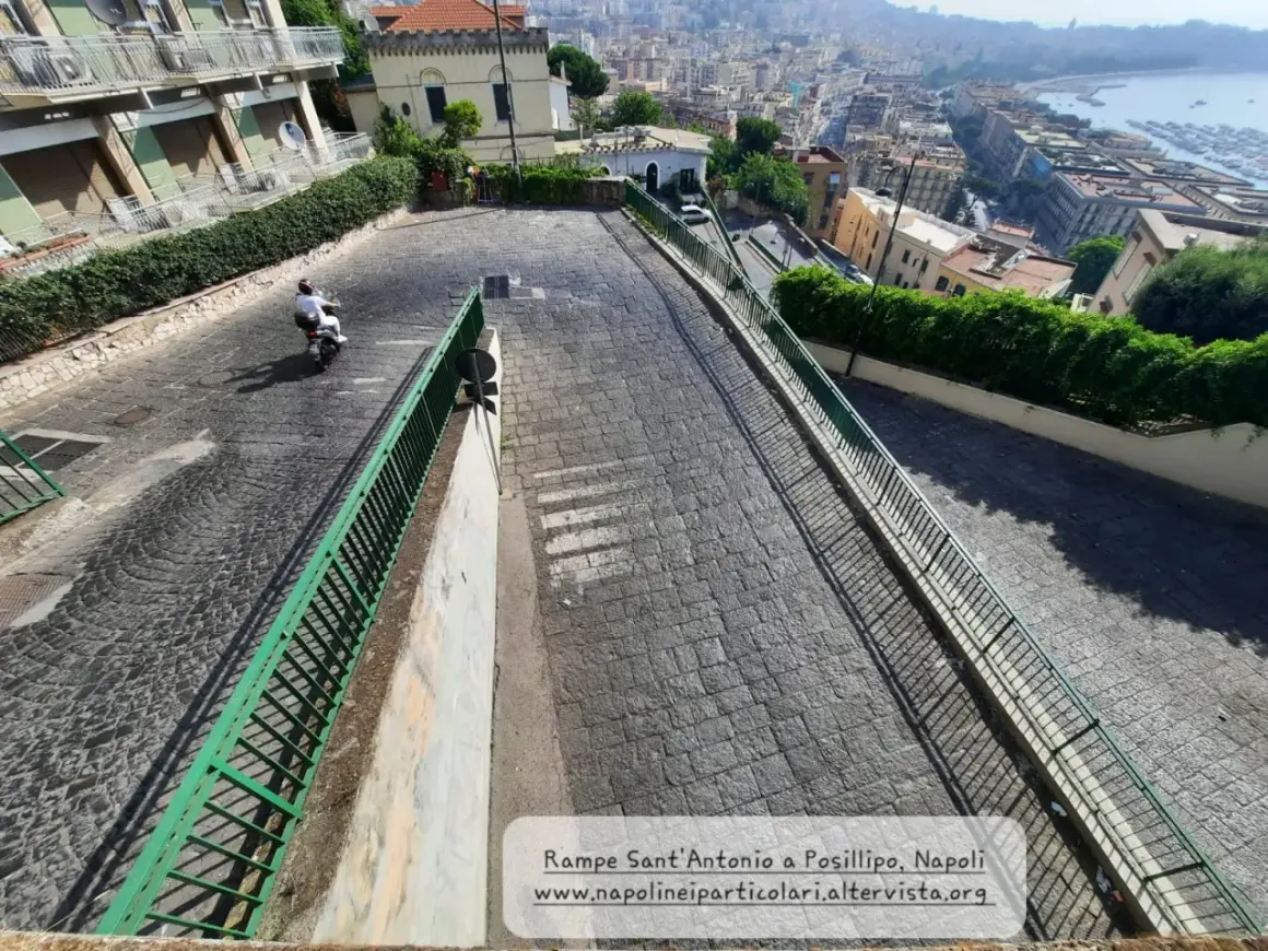 Le 13 discese di Napoli, una vista panoramica di una strada in discesa con un motorino, tra palazzi e vegetazione, che porta verso il mare.
