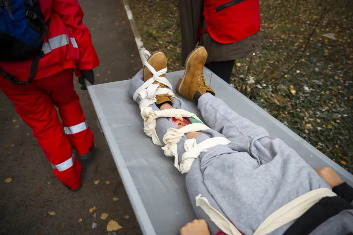 Na noszach leży osoba z opatrzonym złamaniem otwartym nogi. Ratownicy w czerwonych strojach udzielają pomocy.
