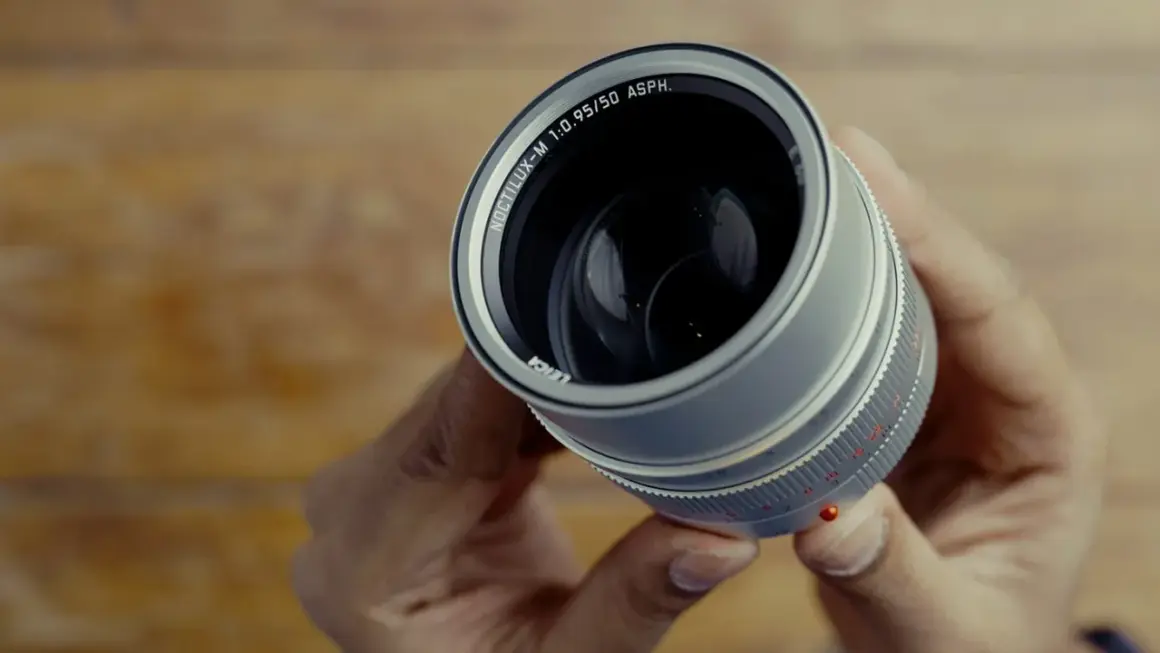 Hands hold a Leica Noctilux M 50 mm lens, its silver barrel and prominent f/0.95 aperture gleaming.