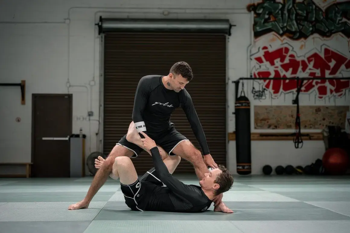 Två män tränar ju jutsu på en matta. Den ena ligger ner medan den andra står över honom, med benen i en låsande position.