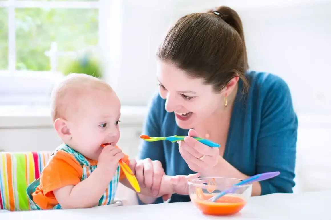 Mama karmi niemowlę, które próbuje samodzielnie jeść. To obrazek z tabeli żywienia 7-miesięcznego dziecka.