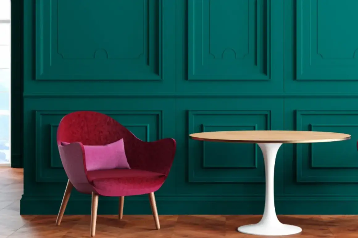 Poltrona rossa vellutata con cuscino rosa, accanto a un tavolino rotondo, su sfondo verde petrolio con pannelli decorativi.