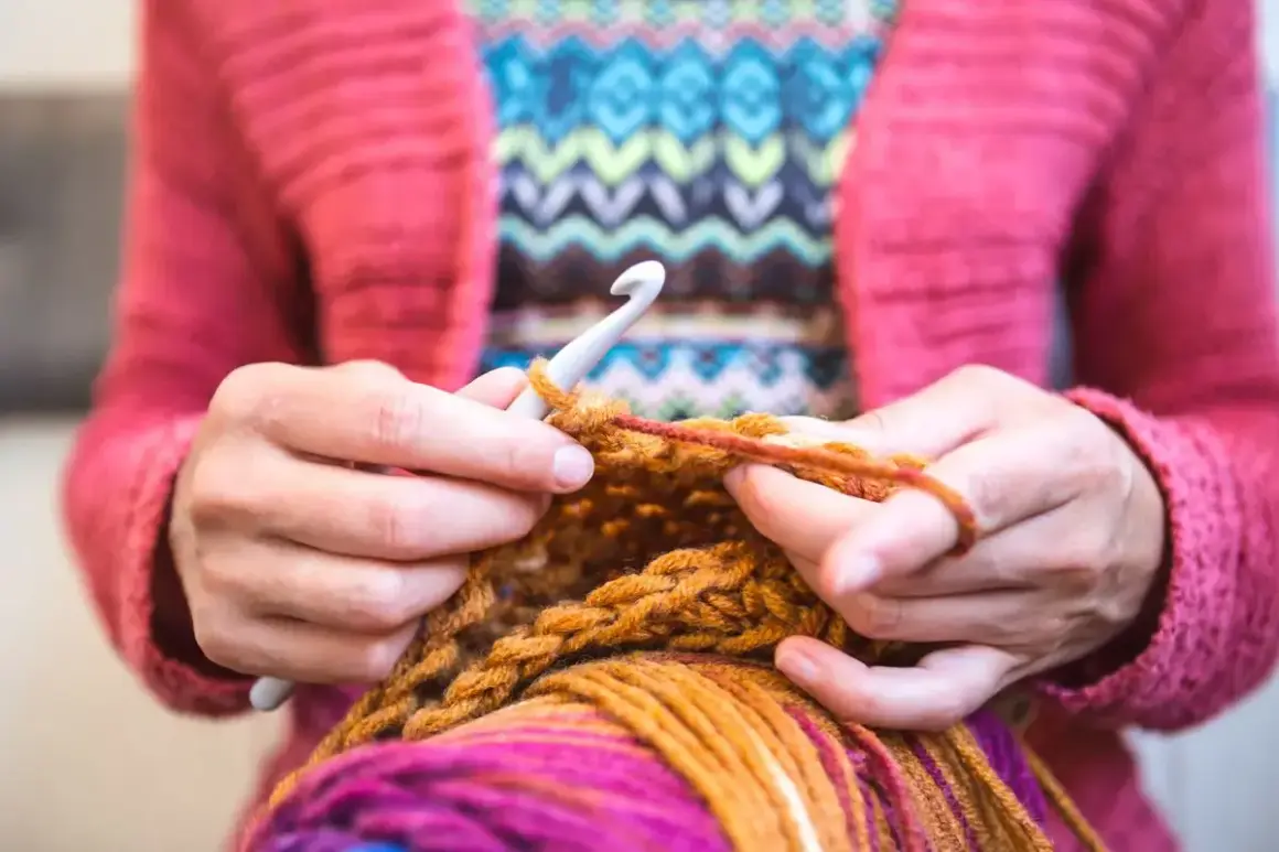 Mani che lavorano a maglia con un uncinetto, filato colorato e un maglione rosa. Nonostante sembri rilassante, lavorare a maglia fa male al cuore se si pensa troppo.