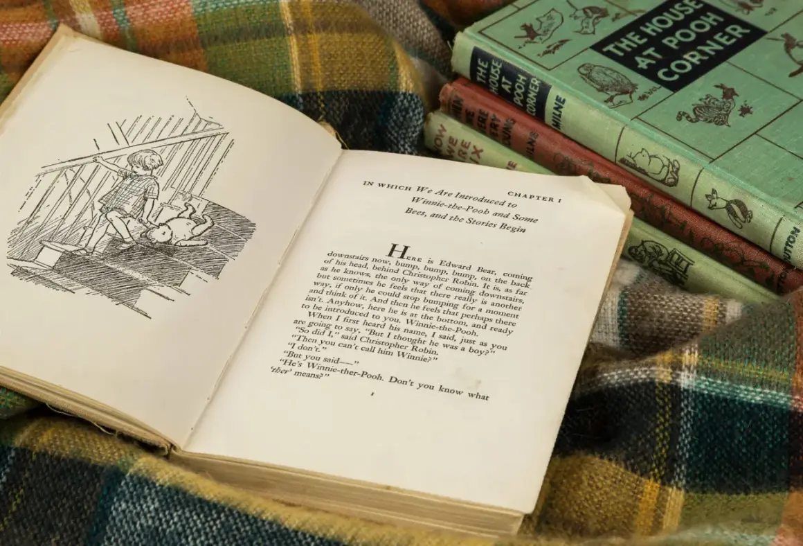 Otwarta książka z ilustracją Kubusia Puchatka i Christophera Robina na schodach. W tle stos książek A.A. Milne'a.