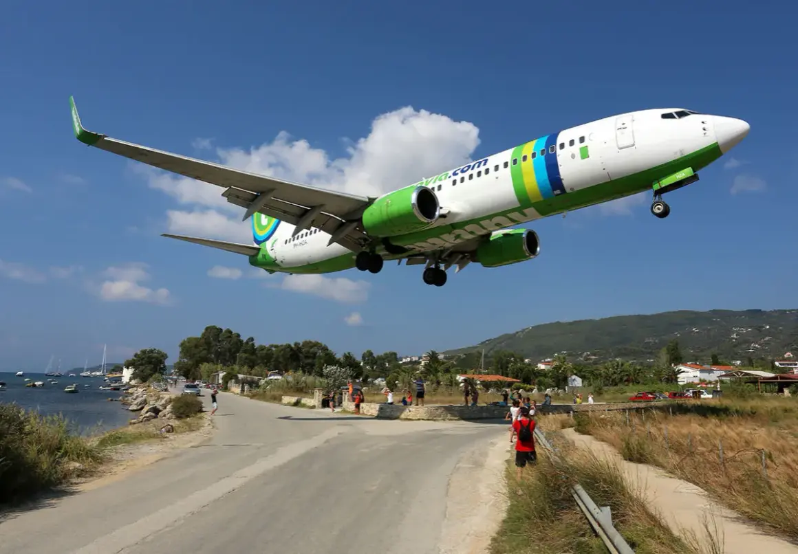 Ett flygplan landar lågt över vägen vid Skiathos flygplats. Människor står längs vägen och tittar upp.