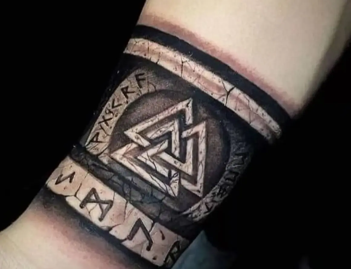 Tatuaże wikingów na przedramieniu: symbol Walhalla i runy, wyglądające jak wyryte na kamieniu.