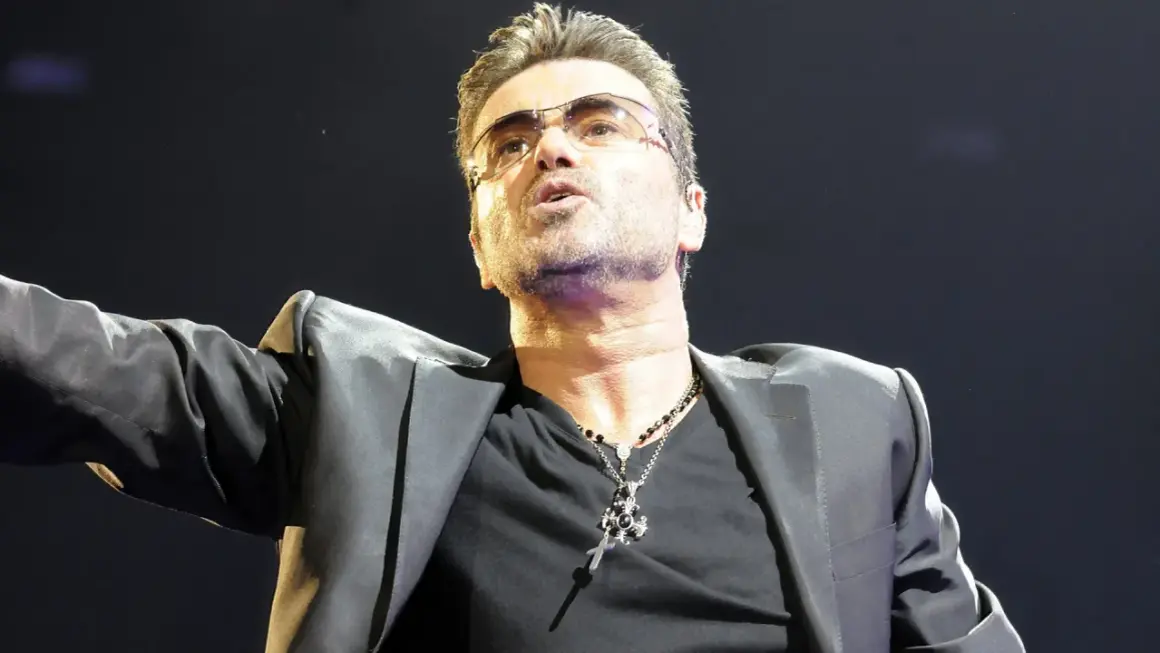 George Michael w okularach, w czarnej marynarce, śpiewa swoje ponadczasowe utwory.