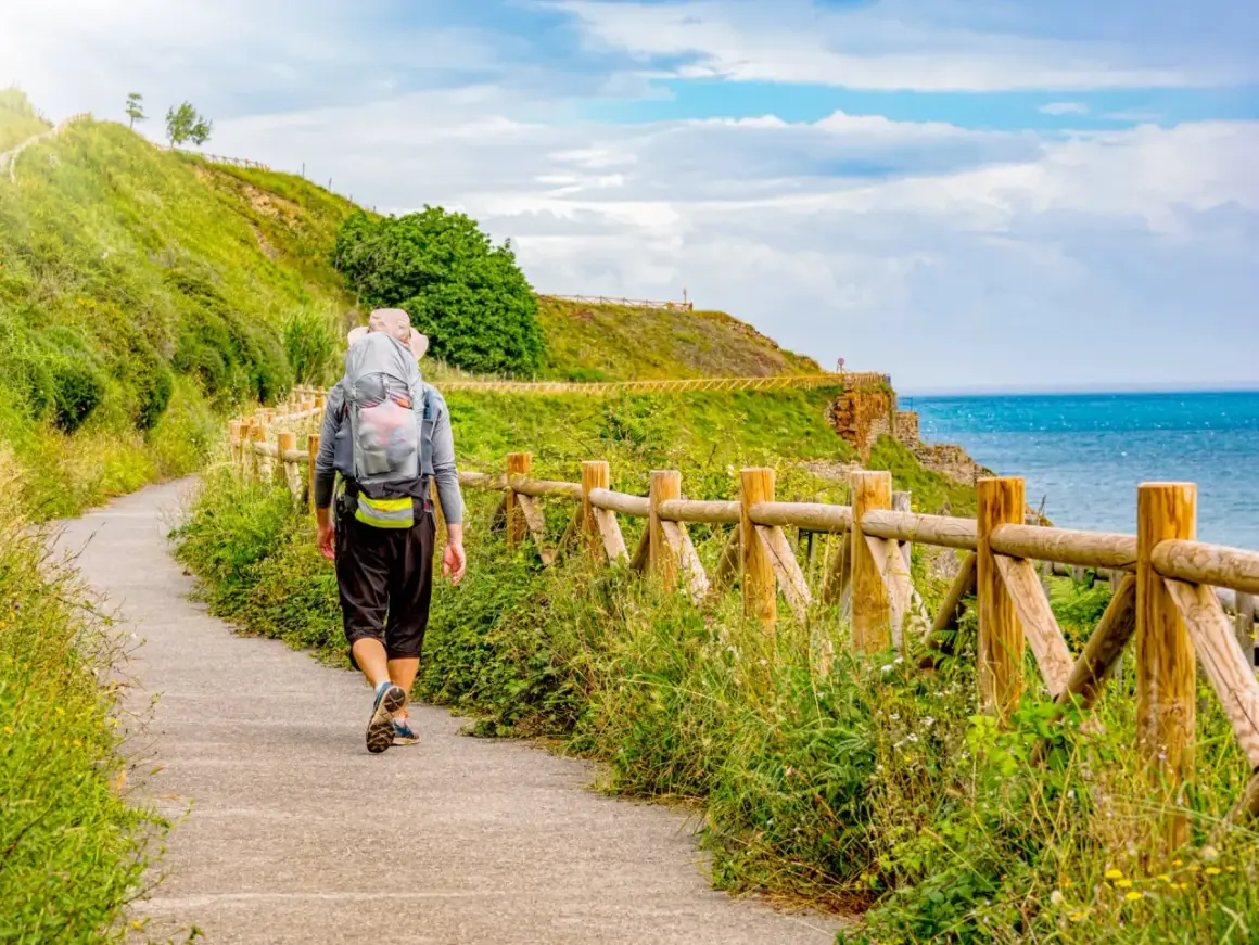 Vandrare på el camino de Santiago längs en kuststig med havet till höger och en grön kulle till vänster.