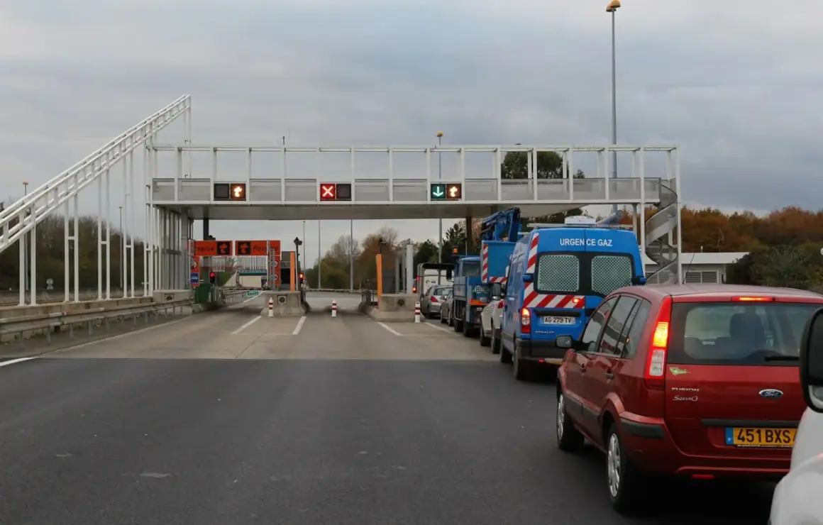 Voiture rouge et camions bleus dans une file d'attente au péage flux libre A13. Lumières rouges et vertes sur les panneaux.