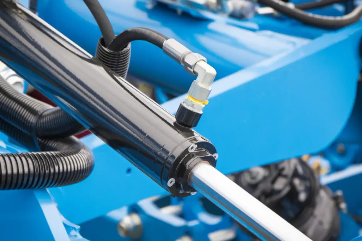 Części do hydroforu: czarny siłownik hydrauliczny z metalowym złączem i wężem, na tle niebieskiej maszyny.