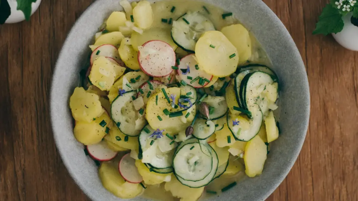 Kartoffelsalat mit Gurken, Radieschen und Schnittlauch, garniert mit kleinen blauen Blüten. Ein frischer Genuss auf Holztisch.
