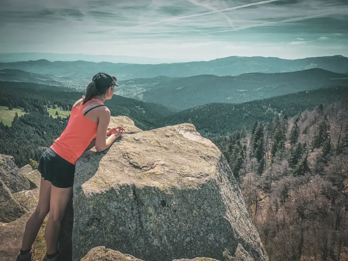 Une femme profite d'une vue panoramique sur les Vosges lors d'un trek. Les montagnes s'étendent à perte de vue sous un ciel nuageux.