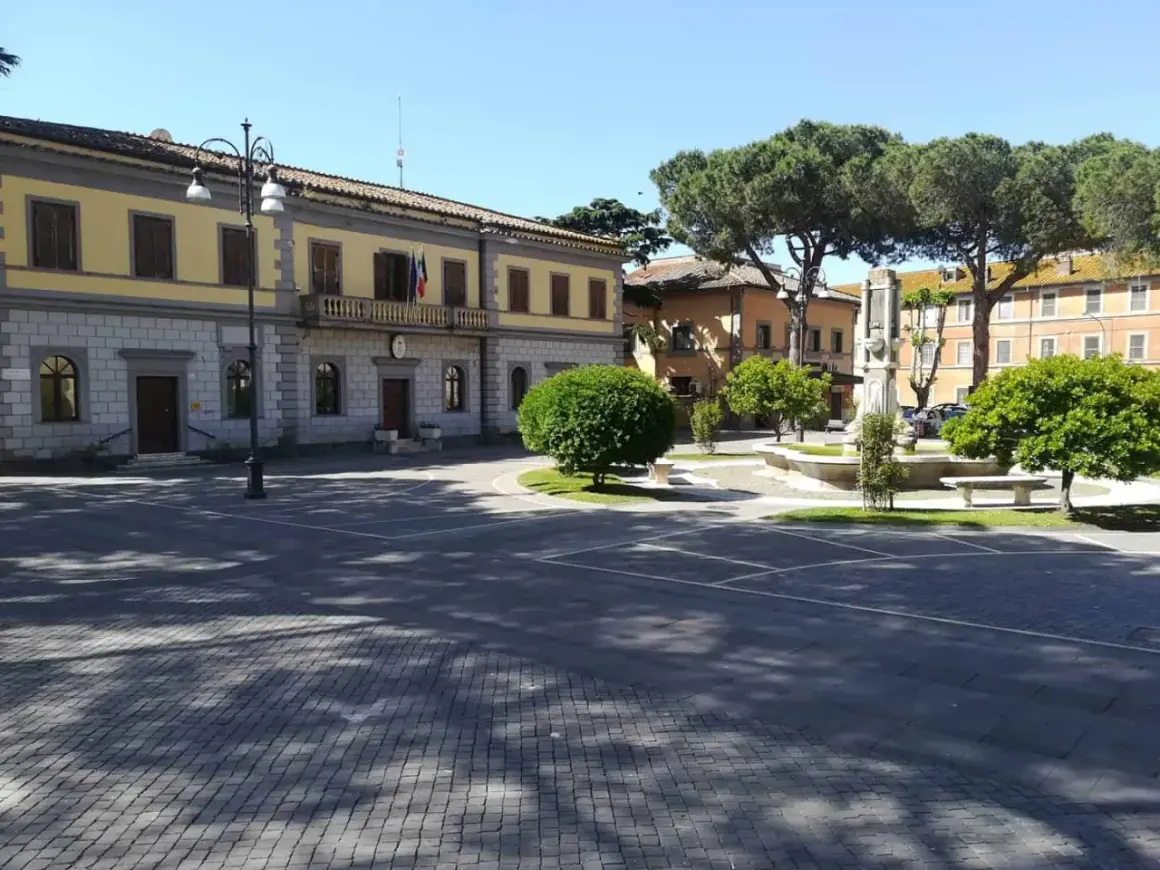 Piazza principale di Monte Romano, Viterbo, con il palazzo comunale, una fontana e alberi rigogliosi sotto un cielo azzurro.