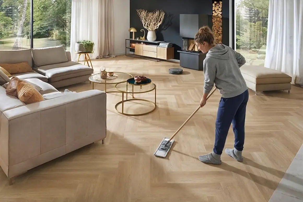Frau wischt den hellen Designboden im Wohnzimmer. Ein Saugroboter ist im Hintergrund zu sehen.