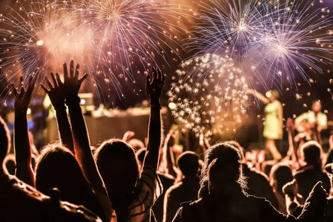 Folla festante al ritmo di canzoni di Capodanno famose, con fuochi d'artificio che illuminano il cielo.