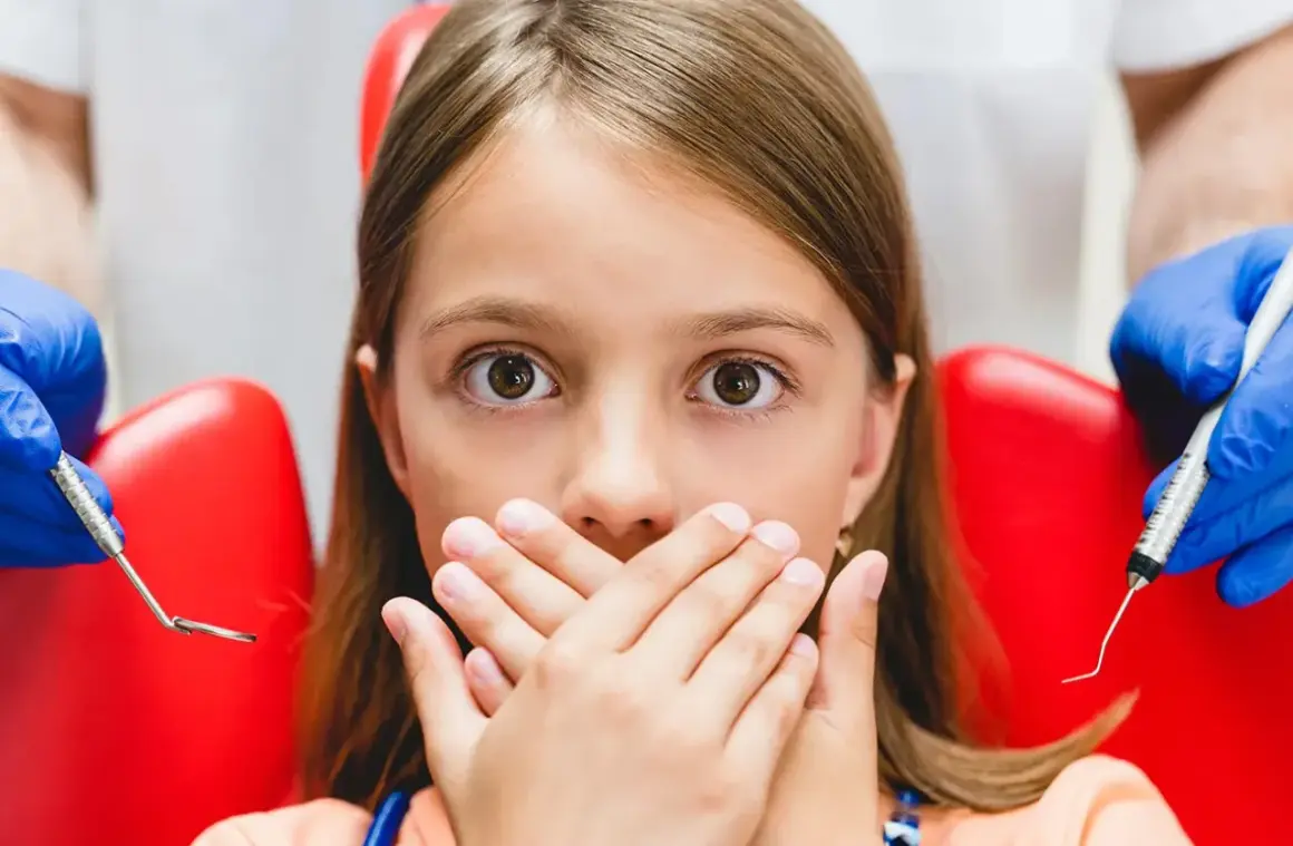 Przerażona dziewczynka zakrywa usta dłońmi, gdy dentysta przygotowuje się do podania znieczulenia. Wokół niej widać czerwony fotel dentystyczny i narzędzia.