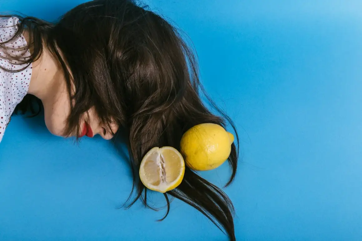En person med mörkt hår och röda läppar ligger på en blå bakgrund. Två citroner, en hel och en delad, ligger bland håret, som om de används för att bleka håret med citron.