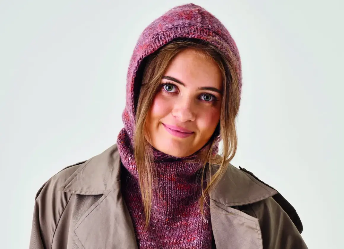 Ung kvinna med blå ögon bär en stickad balaclava i rosa och lila nyanser, med en beige trenchcoat.