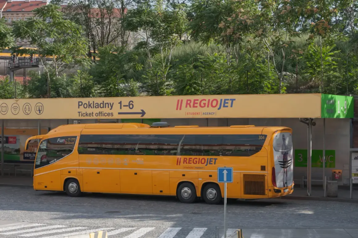 Żółty autobus RegioJet na praskim dworcu. W tle tablica z informacją o kasach biletowych i zielony przystanek.