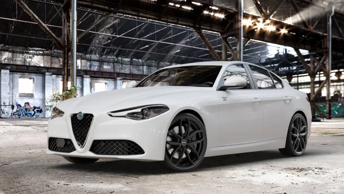 Biała Alfa Romeo Giulia Veloce z czarnymi felgami stoi w opuszczonej hali przemysłowej, gdzie światło przebija się przez dach.