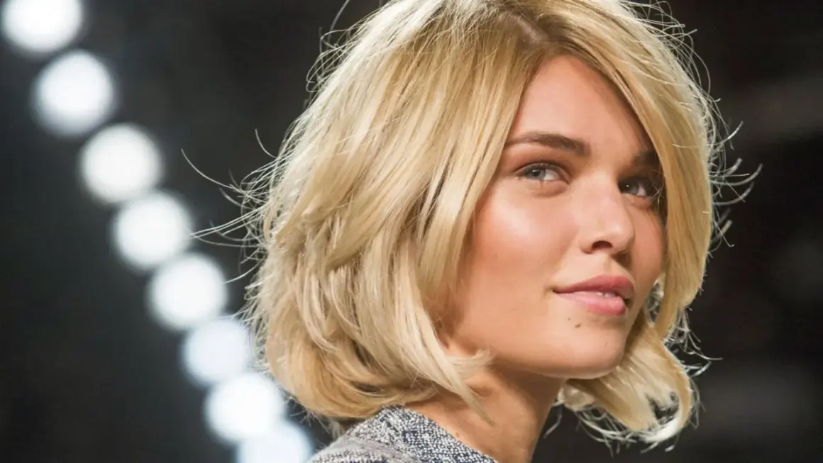 Blonde Frau mit einem stufigen Bob und dicken Haaren, die im Scheinwerferlicht glänzen.