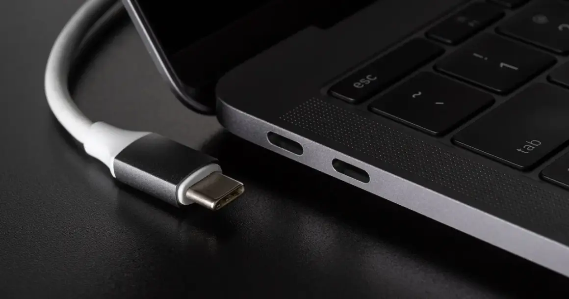 Kabel USB C zbliża się do portu laptopa. Widać jego metalową wtyczkę i biały kabel.