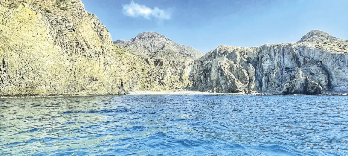 Aguas azules bañan la costa rocosa de una cala en Almería. Montañas escarpadas y una pequeña playa de arena componen el paisaje.