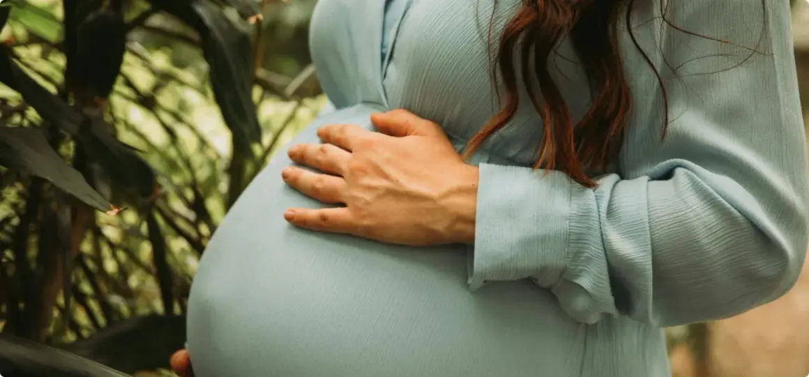 Gravid i vecka 5, en kvinna med ljusblå klänning håller om sin mage bland gröna blad.