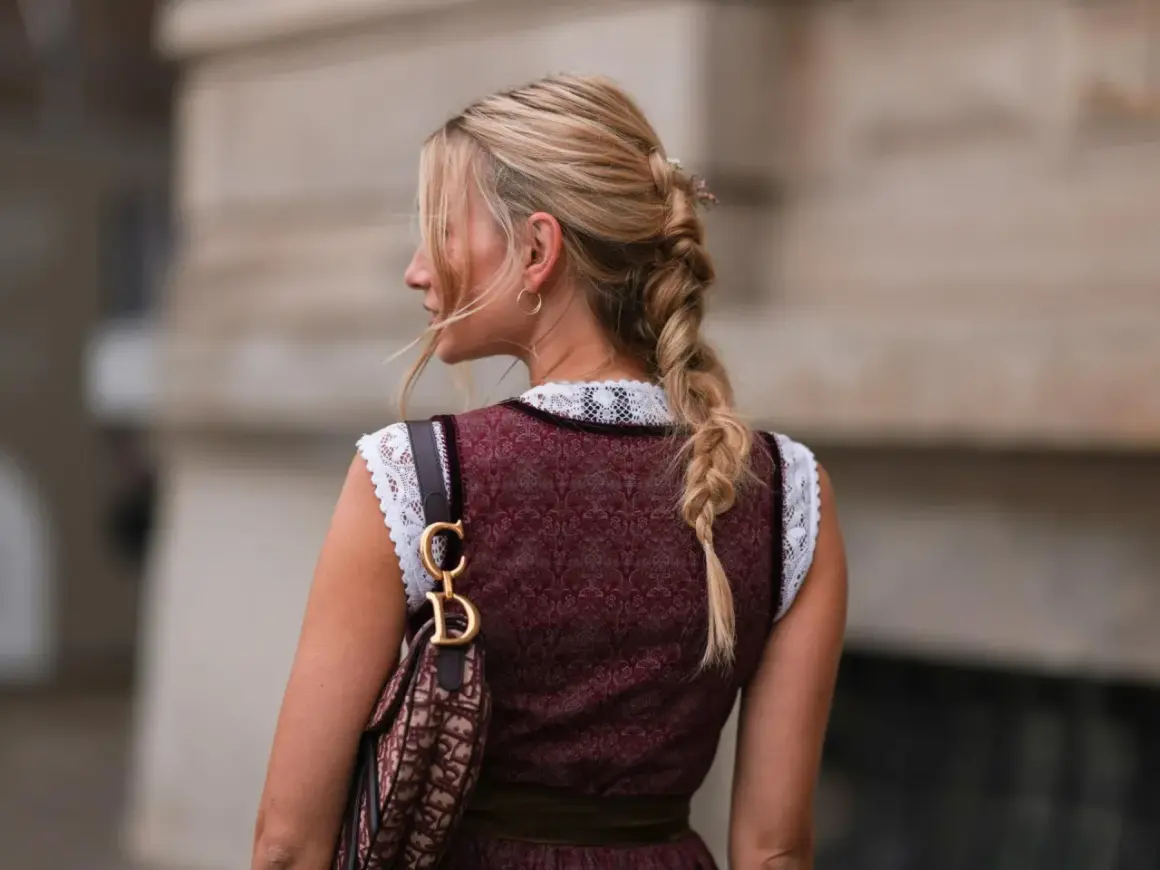 Blonde Zopf-Frisur mit Blumenakzenten, perfekt für Oktoberfest Frisuren. Trägt Dirndl und eine Designer-Tasche.