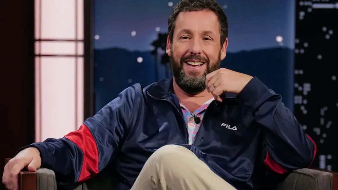 Adam Sandler skrattar glatt i en blå Fila-jacka, redo för en rolig intervju.