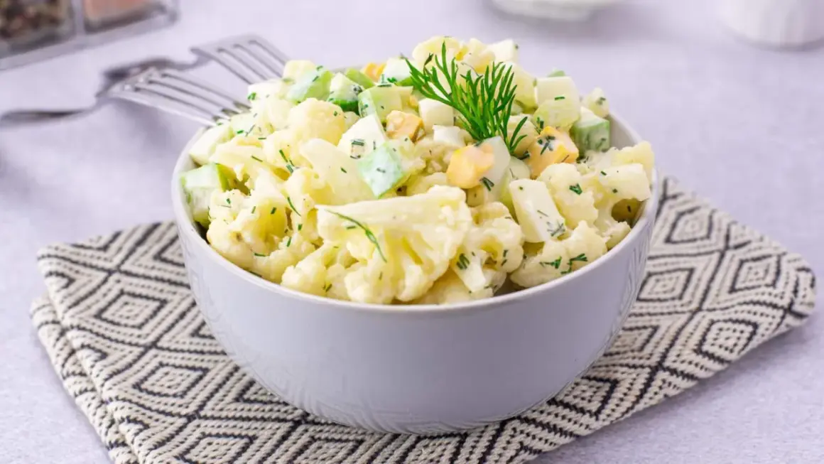 Sałatka z kalafiorem, jajkiem i ogórkiem, udekorowana koperkiem, podana w misce na wzorzystej serwetce.