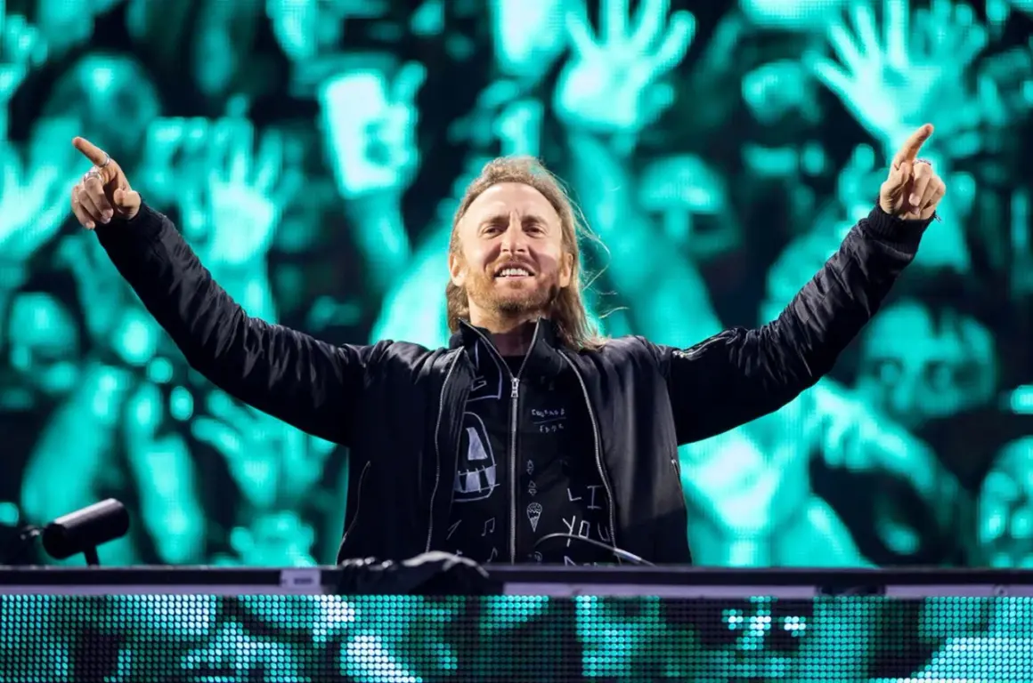 David Guetta na jednym ze swoich koncertów, z rękami uniesionymi w geście radości. Tłum na tle ekranów LED.