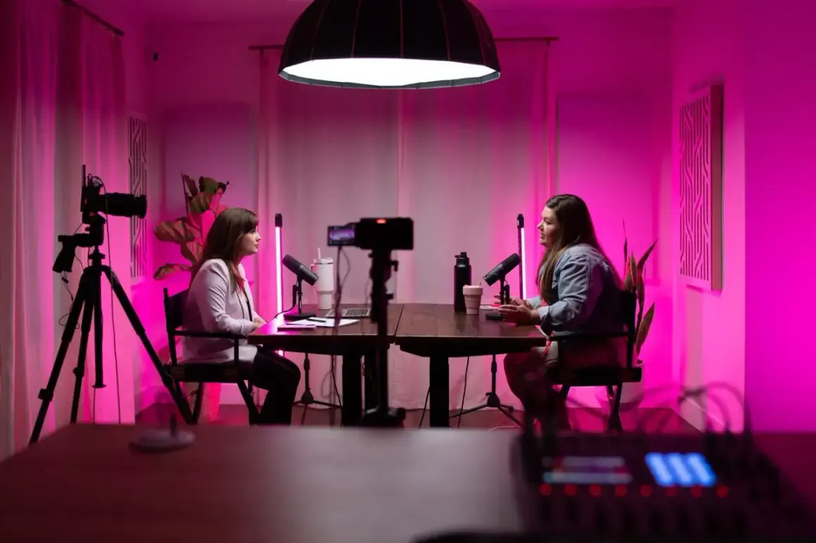 Two women are recording a podcast with a professional podcast lighting setup, featuring pink ambient light and studio microphones.