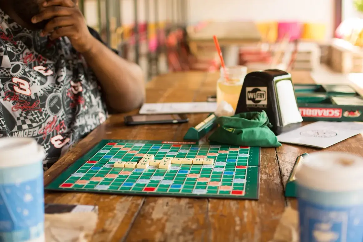 Gra planszowa dla 2 osób, Scrabble, na drewnianym stole. Litery ułożone na planszy, napój i kubek w tle.